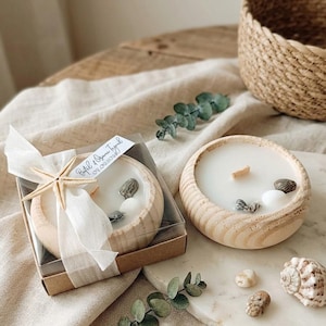 Puede incluir: Dos cuencos redondos de madera con velas de cera blanca y mechas de madera. Una vela est&aacute; en una caja transparente con una cinta blanca y una estrella de mar. Ambas velas tienen conchas marinas. Una etiqueta dice Bet&uuml;l & Oguzhan Toyrul 09.09.2023.