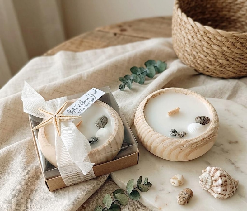 Puede incluir: Dos cuencos redondos de madera con velas de cera blanca y mechas de madera. Una vela est&aacute; en una caja transparente con una cinta blanca y una estrella de mar. Ambas velas tienen conchas marinas. Una etiqueta dice Bet&uuml;l & Oguzhan Toyrul 09.09.2023.