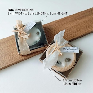 Puede incluir: Dos cajas de regalo cuadradas de 8 cm de ancho, 8 cm de largo y 3 cm de alto, cada una con una vela redonda de cera blanca, conchas peque&ntilde;as y una estrella de mar. Una cinta de lino y algod&oacute;n de 2,5 cm rodea cada caja con una etiqueta.