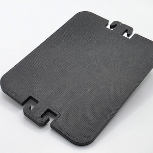 Könnte beinhalten: Ein schwarzes, rechteckiges Objekt mit strukturierter Oberfläche und einzigartigem Design. Das Objekt hat eine abgerundete Form mit drei hervorstehenden Laschen oben und einer einzelnen Lasche unten. Das Objekt besteht aus einem festen, dunklen Material.