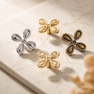 Puede incluir: Cuatro pomos de metal para armarios con forma de flores de cuatro pétalos. Un pomo es de plata pulida, otro de oro cepillado, otro de oro brillante y otro tiene un acabado de bronce antiguo. Cada pomo presenta un detalle cuadrado central.
