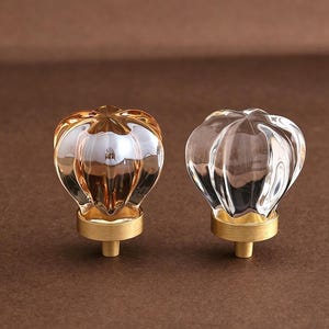 Puede incluir: Dos pomos para armarios con cabezas de cristal facetado en forma de calabaza. Un pomo es de cristal con un tono ámbar cálido, mientras que el otro es de cristal transparente. Ambos están montados sobre bases metálicas de latón cepillado.