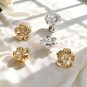 May include: A collection of five metal drawer knobs shaped like flowers. Three knobs are finished in a polished gold tone, while two knobs are finished in a polished silver tone. Each knob features detailed petal designs and a central circular base.