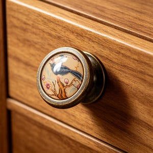 May include: A round cabinet knob with an antique brass finish and a central ceramic insert. The insert features a detailed illustration of a dark blue bird perched on a brown tree branch with light pink flowers against a cream background.