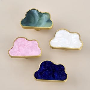 Può includere: Quattro pomelli per armadietto a forma di nuvola con bordi in metallo dorato. Ogni pomello presenta un inserto in resina distinto: uno in verde scuro marmorizzato, uno in bianco solido, uno in rosa chiaro marmorizzato e uno blu con glitter.