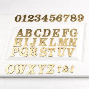 May include: A collection of gold-coloured metal letters, numbers, and symbols. The set includes the digits 0 through 9, the full alphabet from A to Z, an ampersand, an arrow, and a vertical bar, all featuring a clean, serif-style typeface.