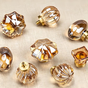 Könnte beinhalten: Eine Sammlung von acht Schrankknöpfen mit bernsteinfarbenen Glasköpfen in verschiedenen facettierten, gerippten und glatten Formen. Jeder Knopf ist an einem gebürsteten goldfarbenen Metallsockel befestigt und zeigt eine Mischung aus runden, quadratischen und sternförmigen Designs.