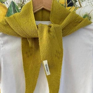 Peut inclure: Un foulard triangulaire en tricot de couleur chartreuse avec un motif texturé et une petite étiquette rectangulaire blanche cousue sur le devant portant l'inscription 100% WOOL. Le foulard est posé sur un t-shirt blanc uni sur un cintre.