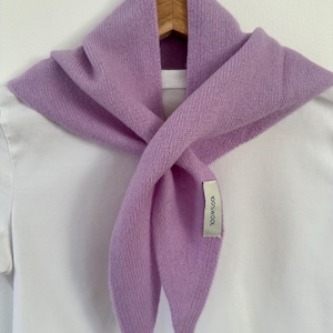 May include: A light purple knit scarf draped over a white t-shirt on a wooden hanger. The scarf features a visible fabric tag on the front that reads 100% WOOL in black text. The knit texture is consistent throughout the triangular-shaped scarf.