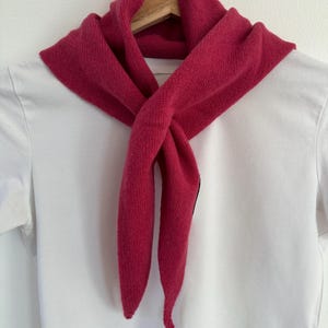 Könnte beinhalten: Ein leuchtend magentafarbener Strickschal, der in einem einfachen Knoten gebunden ist und über einem schlichten weißen T-Shirt liegt. Der Schal hat eine weiche Strickstruktur mit spitz zulaufenden Enden auf dem glatten Stoff.