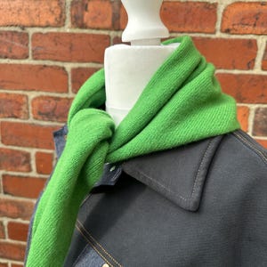 Könnte beinhalten: Ein leuchtend grüner Strickschal, der um den Hals einer weißen Schaufensterpuppe gebunden ist. Der Schal wird über einer dunkel anthrazitfarbenen Jacke mit sichtbaren Nähten und einem strukturierten Kragen getragen. Der Stoff hat eine strukturierte Strickoptik.