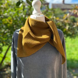 Pode incluir: Um lenço triangular amarelo-mostarda com tecido de malha texturizado, drapeado ao redor do pescoço de um manequim branco. O lenço apresenta um formato pontiagudo e é sobreposto a uma camisa cinza de manga comprida, exibindo um design simples.