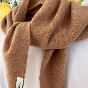 May include: A solid tan-coloured knit scarf made of wool, draped over a wooden hanger. A white rectangular fabric tag is attached to the lower end of the scarf, featuring the black printed text 100% WOOL in a clear, sans-serif font.