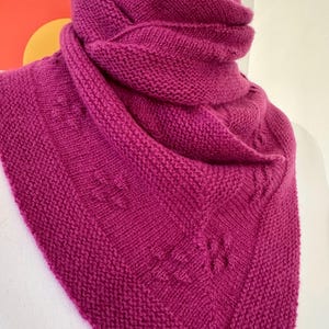 Puede incluir: Un chal triangular de punto color magenta con textura de punto bobo. La prenda presenta un borde ancho con un sutil patrón en relieve similar a hojas a lo largo de los bordes y está drapeado alrededor del cuello en capas.