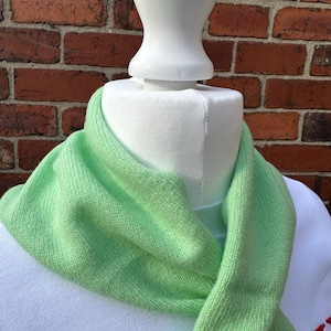 May include: A light lime green knit scarf draped around a white mannequin neck. The scarf features a soft, fine-gauge knitted texture with a consistent colour throughout. The fabric is folded loosely, creating gentle pleats and a smooth, uniform appearance.