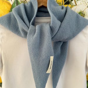 Könnte beinhalten: Ein hellblaues, dreieckiges Stricktuch, das über einem schlichten weißen T-Shirt auf einem Holzkleiderbügel drapiert ist. Das Tuch hat ein sichtbares weißes Stoffetikett an der vorderen Ecke mit dem schwarzen Aufdruck 100% WOOL zur Materialangabe.