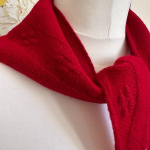 Peut inclure: Une écharpe tricotée rouge vif présentant un motif subtil de points en relief sur tout le tissu. L'écharpe est drapée autour d'un cou de mannequin blanc, mettant en valeur la construction en tricot texturé et la couleur rouge unie du fil.