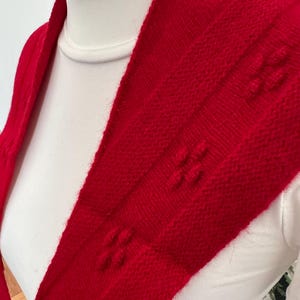 May include: A bright red knitted scarf featuring a textured pattern of small, raised bobble-like clusters arranged in vertical columns. The scarf is draped over a white garment, showcasing the soft, fuzzy texture of the red yarn.
