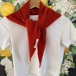 May include: A plain white short-sleeved t-shirt layered with a rust-coloured triangular knit scarf tied at the front. A small white fabric tag is attached to the scarf, featuring the text 100% WOOL printed in black ink on the visible side.