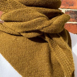 May include: A mustard yellow knitted scarf featuring a textured, chunky stitch pattern. The scarf is wrapped in multiple layers around the neck, displaying a soft, fuzzy yarn texture with a triangular drape that hangs down the front of the garment.
