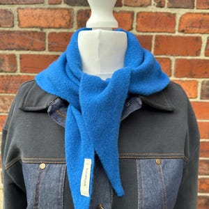 May include: A bright blue triangular wool scarf tied around the neck of a mannequin. The scarf features a white fabric tag with the text 100% WOOL printed in black. The mannequin is wearing a dark grey and blue denim jacket with metal buttons.