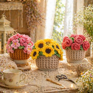 May include: Three crocheted flower arrangements in baskets. One has pink flowers, another yellow sunflowers, and the third red roses. The baskets are brown and beige, sitting on a table with a teacup and scissors.