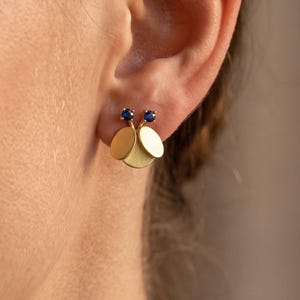 May include: A gold stud earring featuring two circular, matte-finish gold discs layered slightly over each other, topped with two small, round, deep blue sapphire gemstones set in gold prongs, creating a delicate and balanced geometric design.