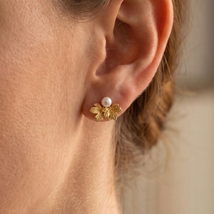 May include: A gold-toned stud earring featuring a textured, multi-lobed leaf design topped with a single, small, round white pearl. The earring is positioned on the earlobe, showcasing a metallic finish and a delicate, organic botanical shape.