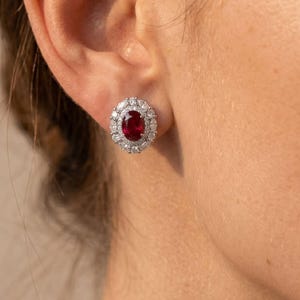 May include: An oval-shaped red ruby stud earring surrounded by a halo of small, clear, round-cut white diamonds set in a polished silver-toned metal frame, worn on the earlobe to showcase the vibrant contrast between the gemstone and diamonds.