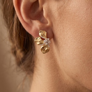 May include: A gold-toned earring featuring three organic, petal-like shapes with a matte finish. The center of the earring is adorned with a cluster of small, clear, sparkling stones that extend slightly along the base of the petals.