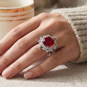 May include: A silver ring featuring a large, cushion-cut red gemstone at the center, surrounded by a halo of small white stones and accented with four small green stones at the cardinal points, set on a band with intricate metal detailing.