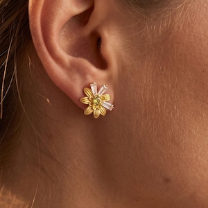 May include: A gold-toned stud earring shaped like a flower with textured petals, a small green center stone, and two clear baguette-cut stones extending from the top, secured to the earlobe with a post and backing mechanism.