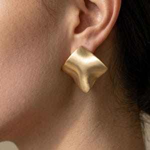Peut inclure: Gros plan d'une boucle d'oreille carrée dorée avec une surface ondulée et texturée. La boucle d'oreille est portée sur l'oreille d'une personne à la peau claire. La boucle d'oreille a une finition mate et un design sculptural unique.