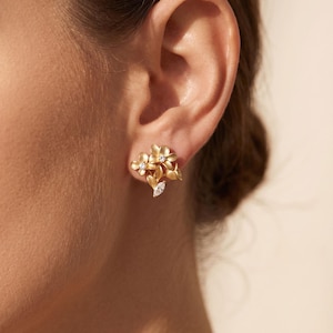May include: A gold-toned stud earring featuring a cluster of three small flowers with matte petals and a single marquise-cut clear stone at the base. The center of each flower is accented with a tiny, sparkling clear round gemstone.