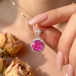 Puede incluir: Un collar de plata con un colgante en forma de rosa rosa. El colgante de la rosa está rodeado por un halo de pequeñas piedras preciosas transparentes. El collar se muestra sobre un fondo borroso.