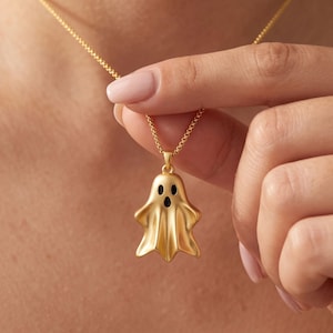 Könnte beinhalten: Eine goldfarbene Halskette mit einem geisterförmigen Anhänger, der eine glatte, fließende Silhouette aufweist und mit zwei schwarzen ovalen Augen sowie einem schwarzen ovalen Mund versehen ist. Der Anhänger hängt an einer feinen Goldkette.