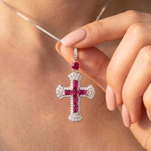 May include: A silver-toned cross pendant necklace featuring baguette-cut red rubies arranged in a cross shape, surrounded by a halo of small round white stones. A single round red ruby sits above the cross, connected to a delicate silver chain.