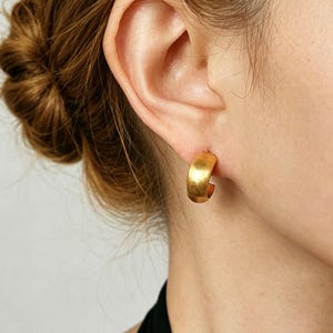 May include: A close-up shot of a gold-coloured hoop earring. The earring is a thick, rounded shape and is worn on the ear. The earring has a matte finish and reflects light. The background is blurred, focusing attention on the jewellery.