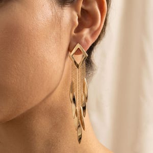 Puede incluir: Un pendiente colgante de tono dorado con un poste superior en forma de diamante conectado a varias cadenas verticales delicadas. Cada cadena termina en un colgante de metal liso, alargado y con forma de hoja, creando un efecto de flecos en cascada.