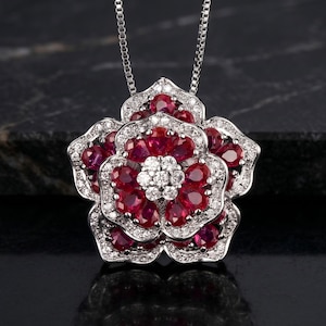 Könnte beinhalten: Eine Halskette mit einem floralen Anhänger in Form einer Blume. Der Anhänger ist aus Silber gefertigt und mit roten Edelsteinen und weißen Diamanten besetzt. Die Halskette wird auf dunklem Untergrund präsentiert.