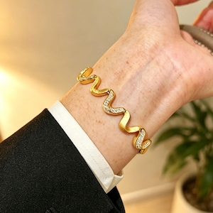 Pode incluir: Uma pulseira dourada com um design ondulado, adornada com cristais brilhantes. A pulseira é usada no pulso, combinada com um blazer preto e uma camisa branca. A joia adiciona um toque de elegância.