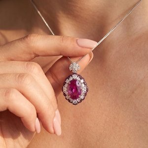 May include: A silver necklace featuring an oval-cut vibrant pink gemstone pendant. The center stone is surrounded by a halo of small white diamonds and a secondary outer border of tiny dark pink stones, all set in a scalloped metal frame.
