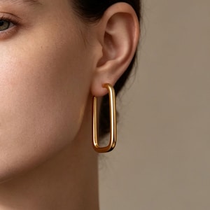 May include: Close-up of a gold-coloured, rectangular hoop earring. The earring has a simple, modern design with a polished finish. The earring is worn on the ear of a person with fair skin, with a neutral background.