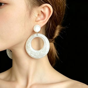 May include: White, large hoop earrings with a textured design and a smaller, round stud. The earrings are a bright white color and have a glossy finish. The earrings are a statement piece.