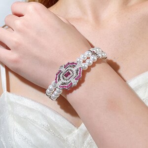May include: A pearl bracelet with a central decorative element. The centerpiece features a rectangular ruby surrounded by white gemstones and pink accents. The bracelet is composed of two strands of white pearls.