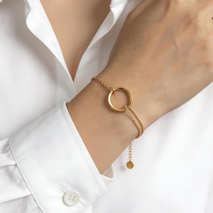 Könnte beinhalten: Ein goldfarbenes Armband mit einem großen kreisförmigen Anhänger. Das Armband besteht aus feinen Kettengliedern und hat einen kleinen, runden Anhänger am verstellbaren Verschluss. Der Schmuck wird an einem Handgelenk, vor einem weißen Hemd getragen.