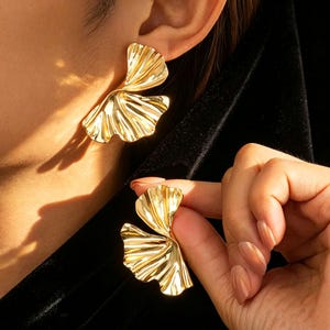 May include: Gold-coloured statement earrings with a layered, fan-like design. The earrings have a textured surface and a shiny finish. One earring is worn on an ear, while the other is held by a hand.