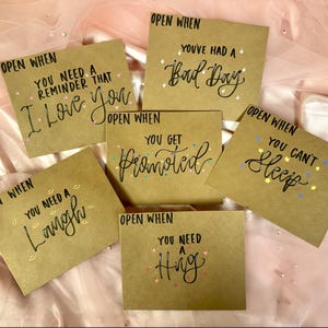May include: Six kraft paper cards with handwritten messages in black ink. Each card is labelled "Open When" followed by a different sentiment, such as "You've Had a Bad Day" and "You Can't Sleep". The cards are displayed on a pink fabric surface.