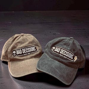Puede incluir: Dos gorras de béisbol en color tostado y gris carbón, cada una con un parche bordado que muestra un icono de calavera, un rayo y el texto "BAD DECISIONS DISCO DIRT" en letras blancas sobre un fondo negro con un borde blanco.