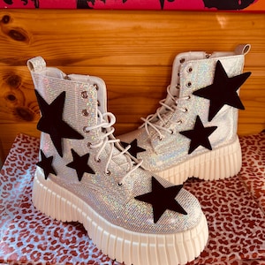 Puede incluir: Botas de combate plateadas con purpurina y aplicaciones de estrellas negras. Las botas tienen cordones blancos, una suela de plataforma acanalada y cierre de cremallera. Una pieza de moda divertida y moderna.